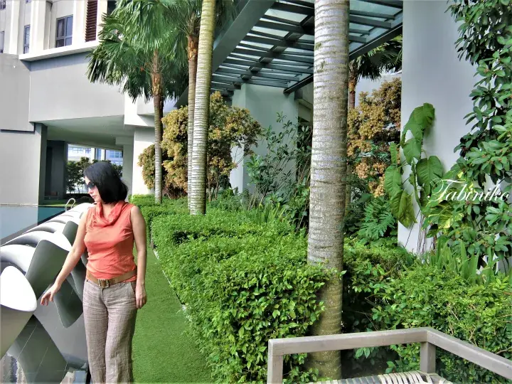 Woman in orange top and beige pants standing by a poolside with modern lounge chairs and tropical greenery at a Malaysia property.