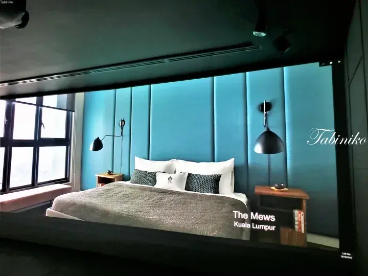 The Mews Kuala Lumpur bedroom with teal padded wall, modern black lamps, and large window view.