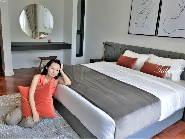Woman in coral top sitting on a rug beside a modern hotel bed with grey and orange accents.