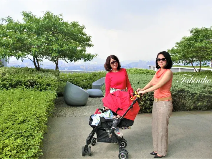Two women push a baby stroller on a garden terrace with modern seating and a view of the city skyline.