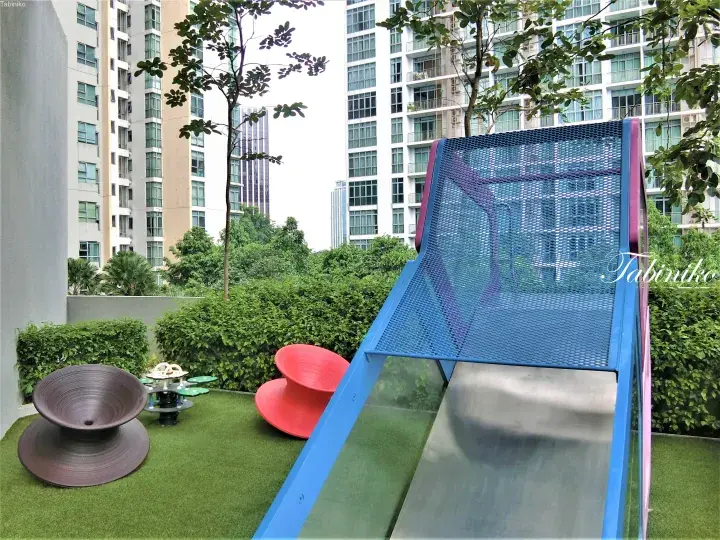 Modern playground with blue slide and colorful spinning seats on artificial grass, surrounded by high-rise condos and greenery.