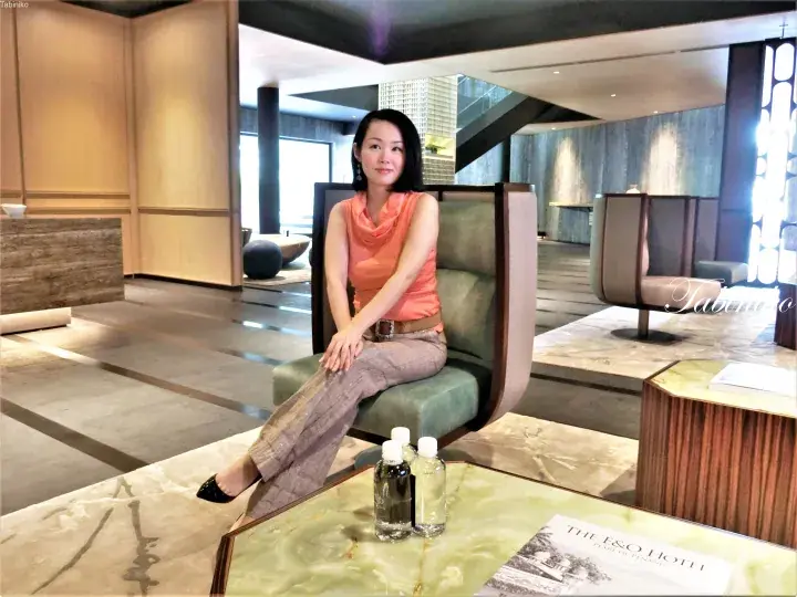 Woman in orange top and grey trousers sitting in a modern lounge chair at The E&O Hotel lobby.