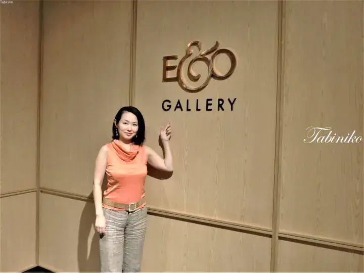 Woman in orange top pointing at the E&O Gallery sign on a wooden wall.