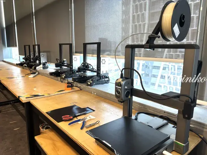 Multiple Creality 3D printers lined up on a wooden table near a window in a modern workspace.