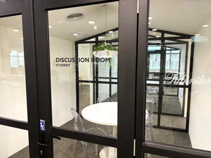 Glass door entrance to a modern student discussion room with a white table and transparent chairs.