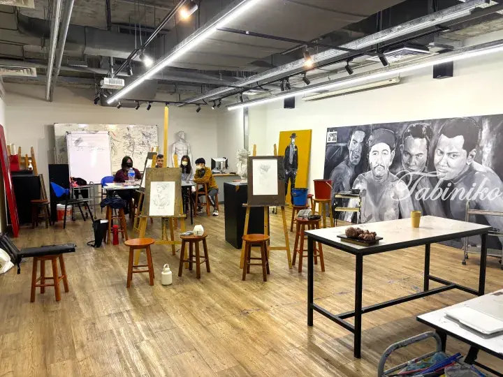 Students sketching plaster busts on easels in a bright art studio with large murals and wooden floors.