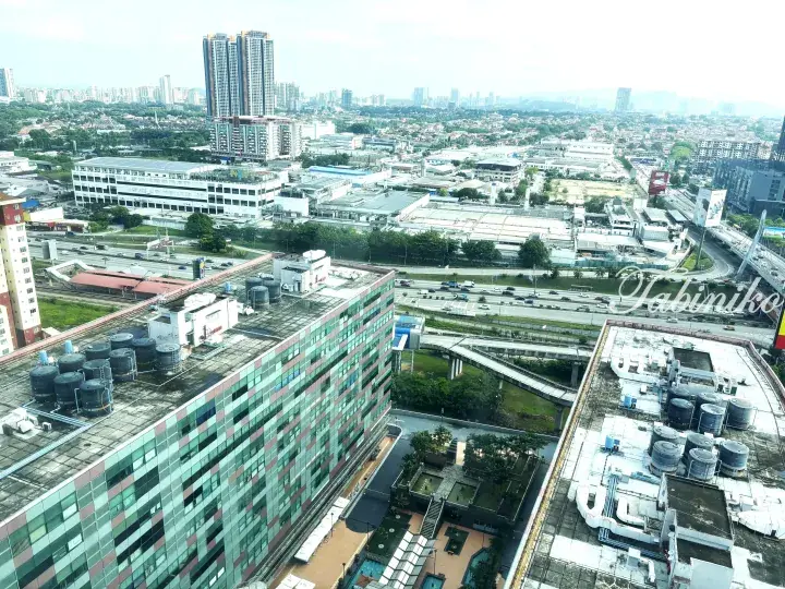 Aerial view of a Malaysian city showing a multi-story building with rooftop water tanks, a highway, and distant high-rise towers.