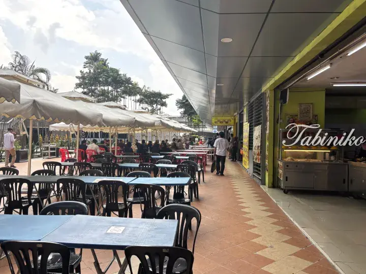 Outdoor dining area with blue tables and beige umbrellas at a Malaysian food court.