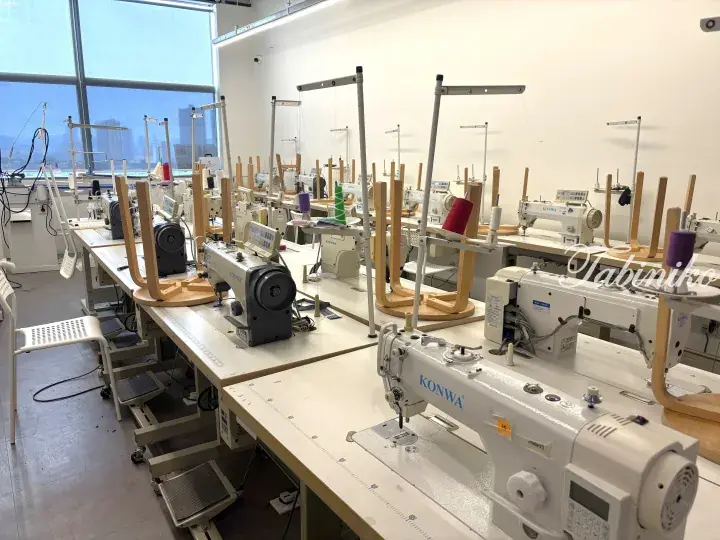 Rows of industrial sewing machines in a bright, modern garment workshop with large windows overlooking the city.