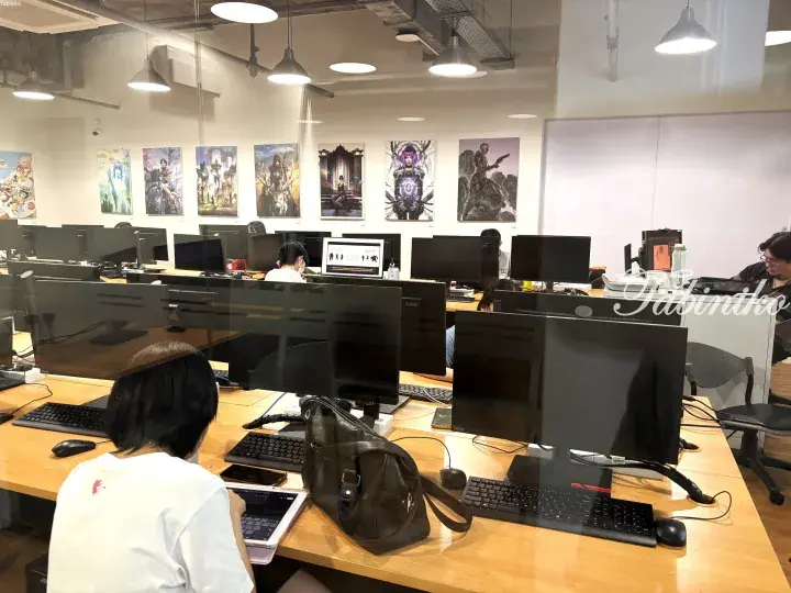 Open-plan office with employees working at desks, featuring multiple monitors and fantasy art posters on the walls.