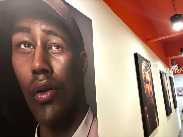 Large portrait of a man displayed on a white wall in a gallery with orange ceiling and other framed artworks.