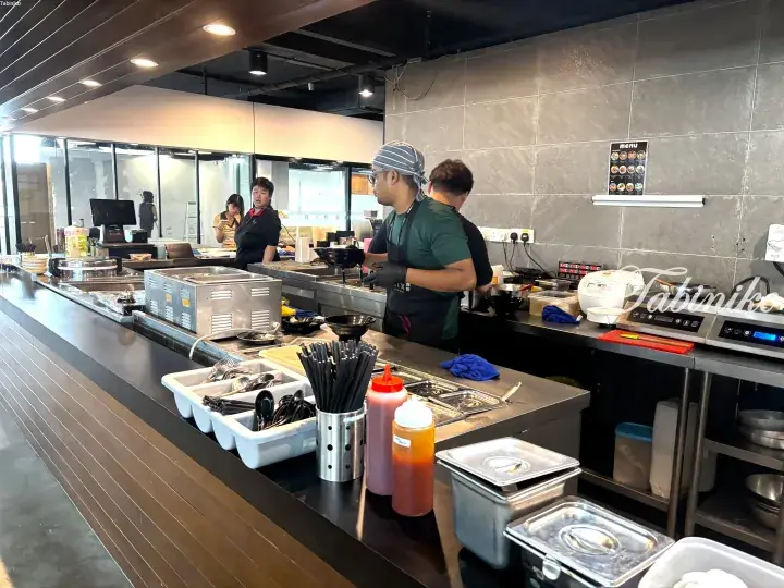Staff preparing food in a modern open kitchen with stainless steel counters and condiments.