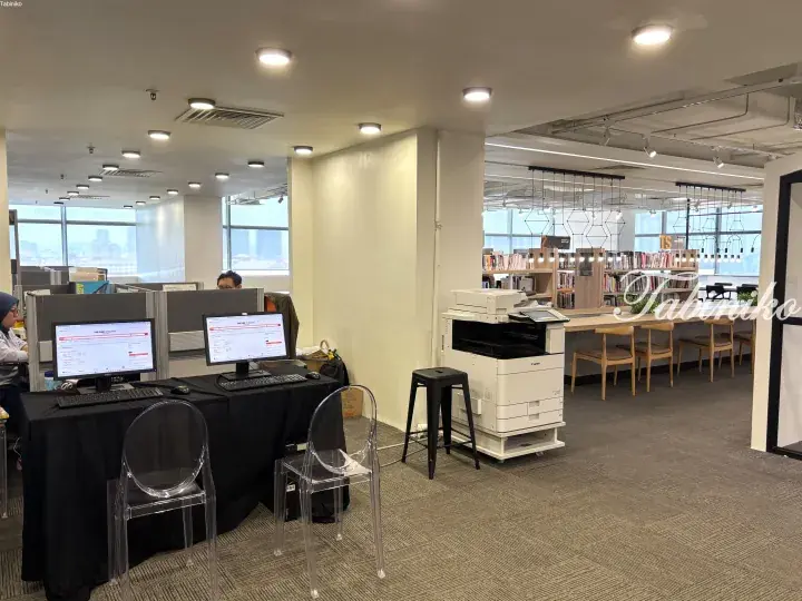 Modern library interior with computer workstations, a large printer, and bookshelves near windows with city views.