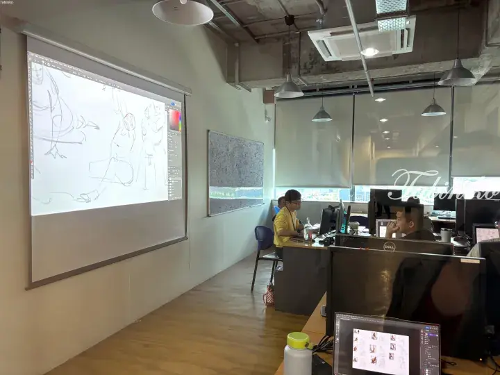 Digital artists work at computers in a modern office while an animation sketch is projected on a large screen.