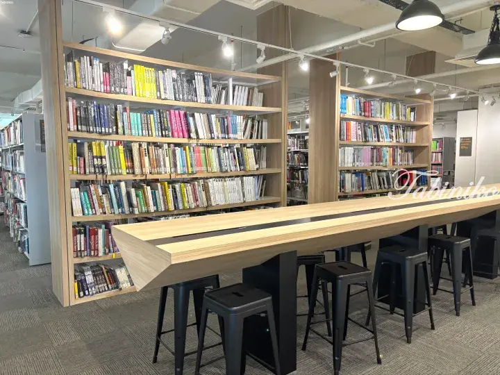 Modern library reading area with wooden bookshelves, long study table, and black stools in a bright interior space.