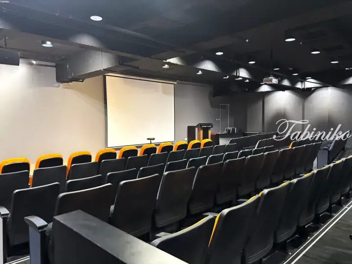 Modern lecture hall with tiered seating, orange and black chairs, and a large projection screen at the front.