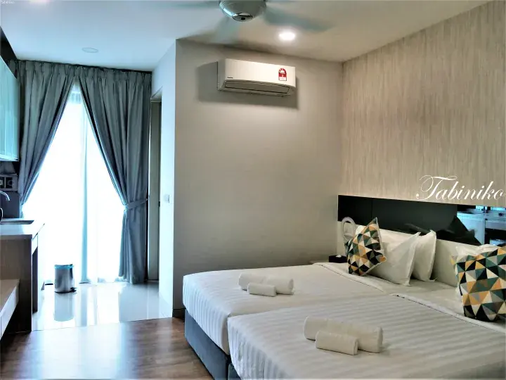 Bright hotel room with two neatly made beds, modern decor, and large window with grey curtains.