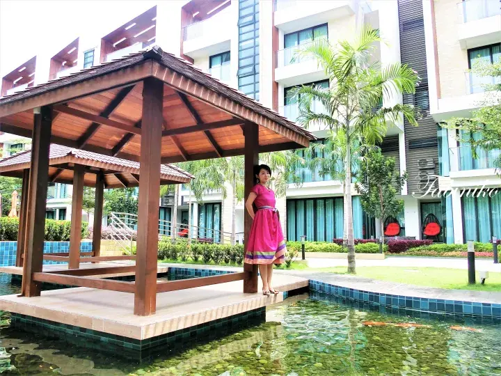 Woman in pink dress standing by wooden pavilion at resort pool with modern building and koi pond.