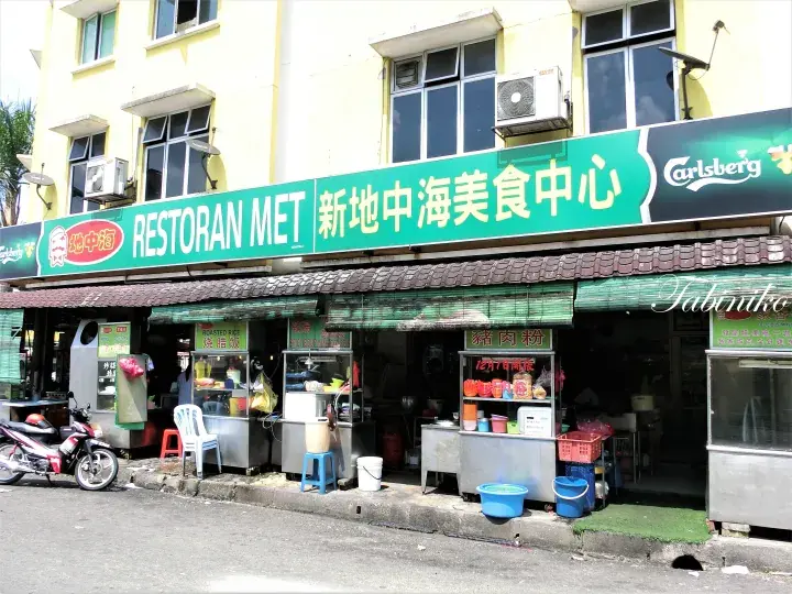 Restoran MET Xin Di Zhong Hai Food Centre storefront with green sign, food stalls, and parked motorcycle.