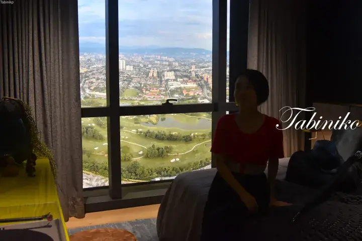 Woman in red top sits on a bed in a high-rise room overlooking a lush golf course and city skyline.