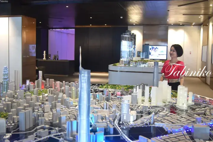 Woman in red top standing beside a detailed architectural model of a modern city skyline featuring a tall skyscraper.