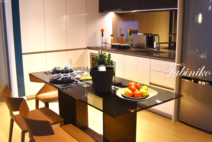 Modern kitchen with a black glass dining table, wine, and fruit, featuring sleek white cabinetry and stainless steel appliances.