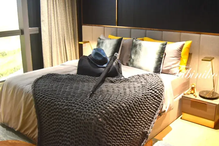 Modern hotel bedroom with a bed featuring a grey knit throw, yellow pillows, and a suitcase with a hat.
