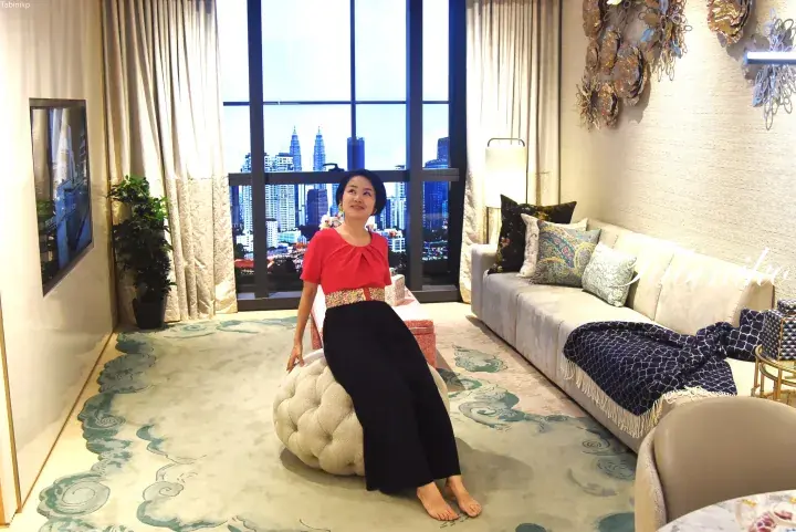 Woman in red top and black skirt sitting on a tufted ottoman in a luxury showroom with a view of the Petronas Twin Towers.