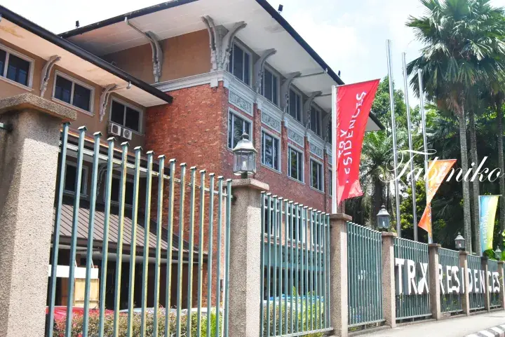TRX Residences exterior showing brick facade, green fence, and red flags with palm trees in the background.