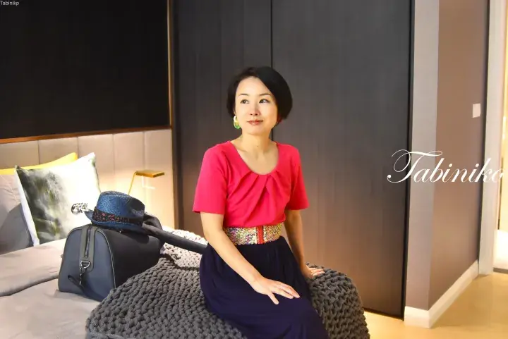 Woman in a pink top and navy skirt sitting on a bed in a modern hotel room with a suitcase and hat nearby.