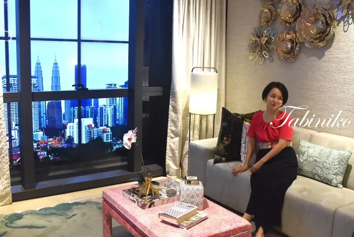 Woman in red top and black skirt sits on a sofa in a luxury apartment, with a view of the Petronas Twin Towers through the window.