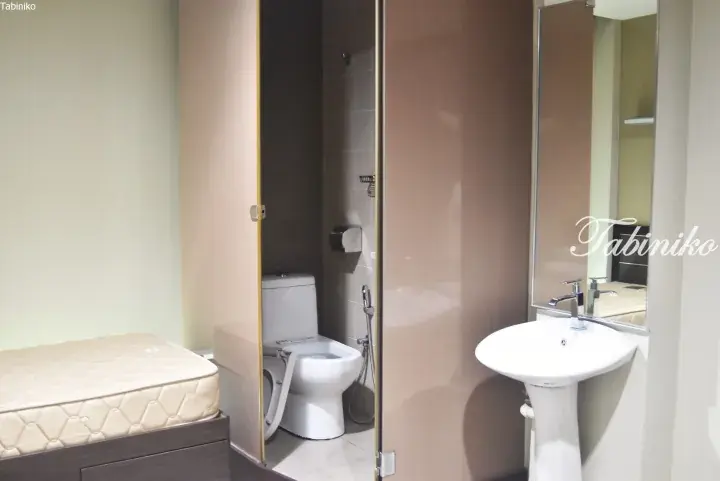 Compact hotel room featuring a bed, a separate toilet cubicle with a bidet, and a pedestal sink with a mirror.