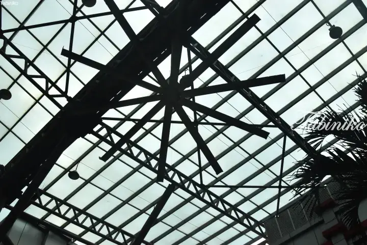 Low-angle view of a large industrial ceiling fan beneath a glass skylight roof with steel beams.