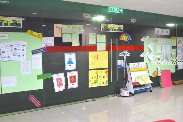 Classroom display with student artwork and bilingual signs on glass walls, featuring a ‘KELUAR’ exit sign.