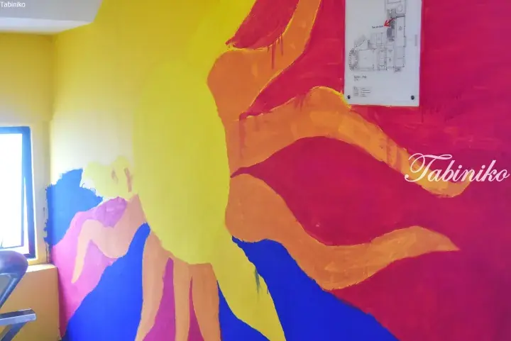 Colorful abstract mural on a wall featuring a large yellow sun, with a floor plan map of Block J Level 1 displayed nearby.