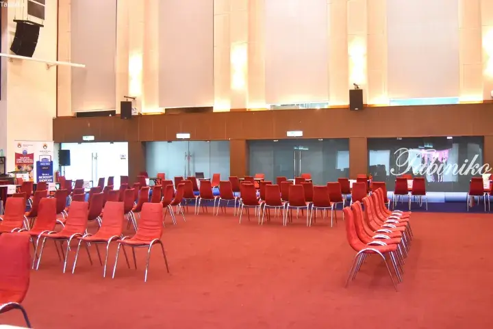 Empty conference hall with red chairs arranged in rows on a red carpet, featuring a stage area and large projection screens.