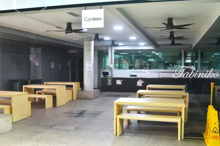 Empty canteen with yellow tables and benches, ceiling fans, and a food counter behind glass.