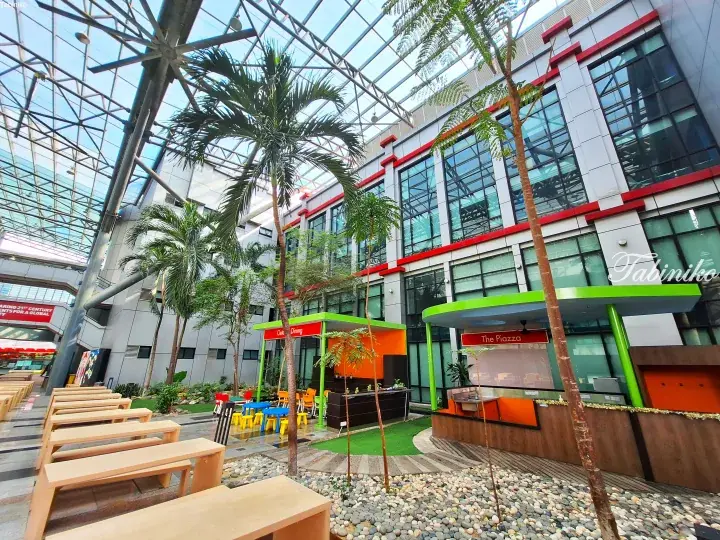 Modern atrium with palm trees, wooden benches, and food stalls under a glass roof.