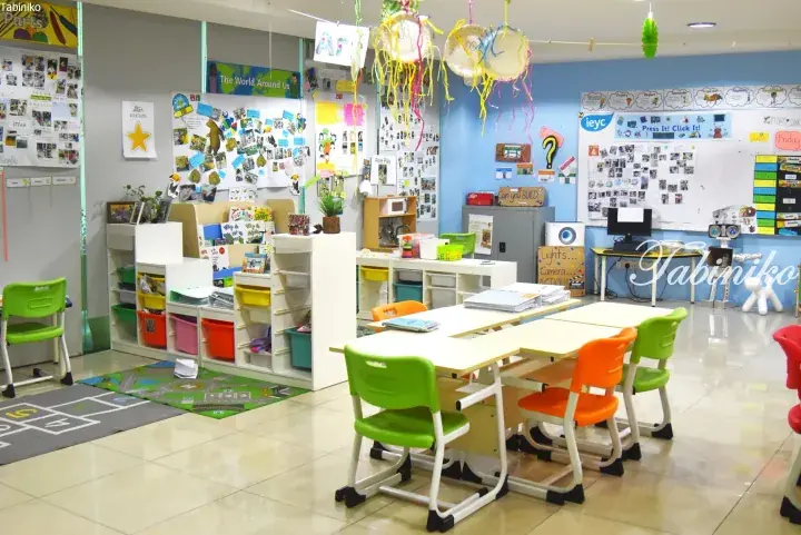 Bright preschool classroom with colorful tables, chairs, and educational displays.