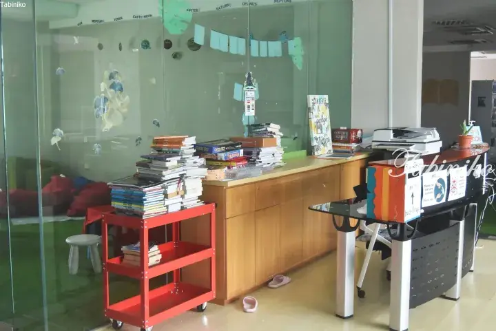 Stacks of books on a red cart and wooden counter in a bright library with glass walls and solar system decorations.