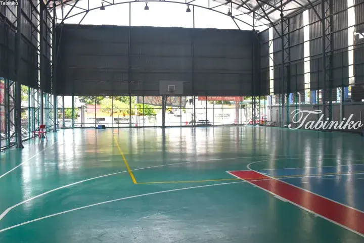 Spacious indoor basketball court with green flooring and red and blue painted areas, featuring a high metal roof structure.