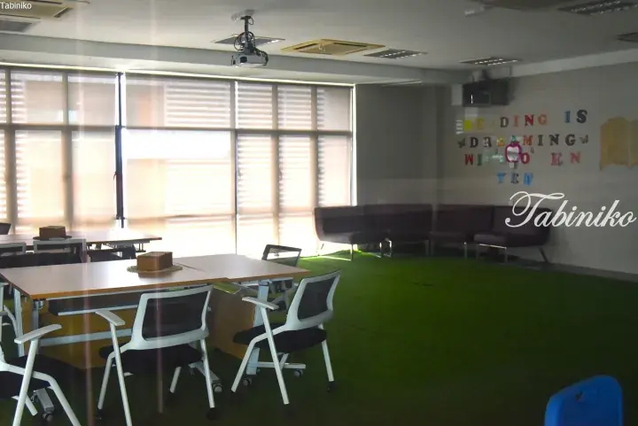 Bright classroom with green carpet, desks, chairs, and a wall quote about reading.