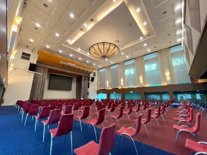 Spacious conference hall with red chairs, blue carpet, and a large screen at the front.