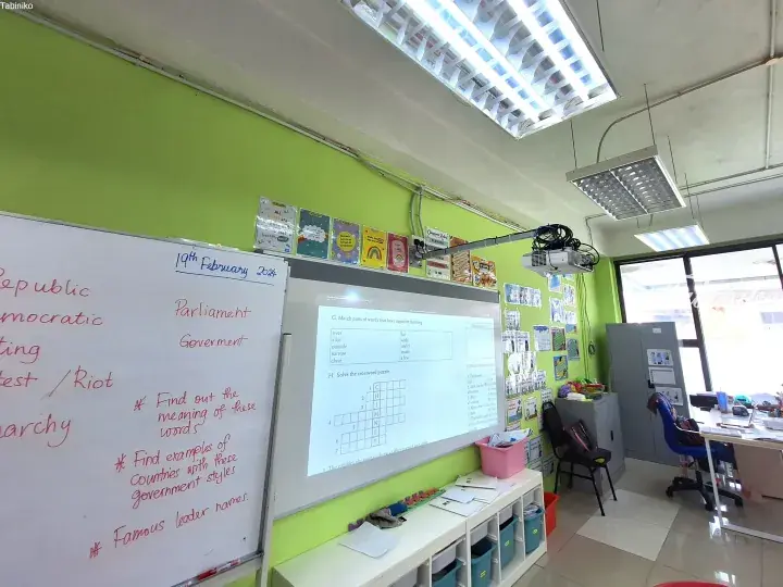 Classroom with green walls showing a whiteboard with ‘19th February 2024’ and an interactive screen displaying English exercises.
