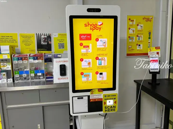 Self-checkout kiosk at PANAS Express convenience store in Malaysia, displaying yellow ‘shop & pay’ instructions for scanning items and e-wallet payments.