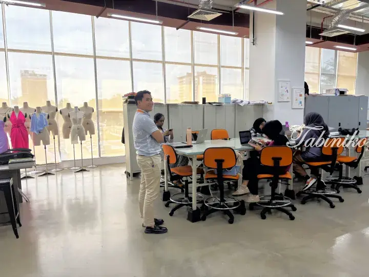 Man standing in a bright fashion design studio with mannequins and students working at tables near large windows.