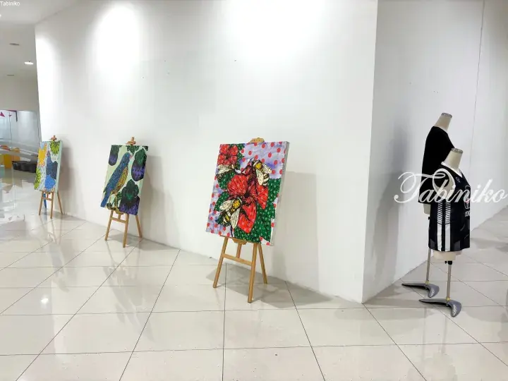 Colorful paintings of birds and flowers displayed on wooden easels alongside mannequins in a bright gallery space.