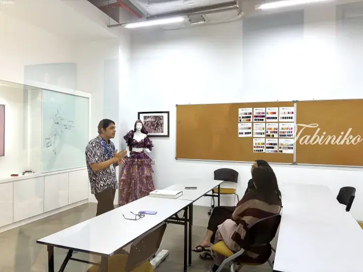 A man in a batik shirt presents a purple outfit on a mannequin to two women in a design studio.
