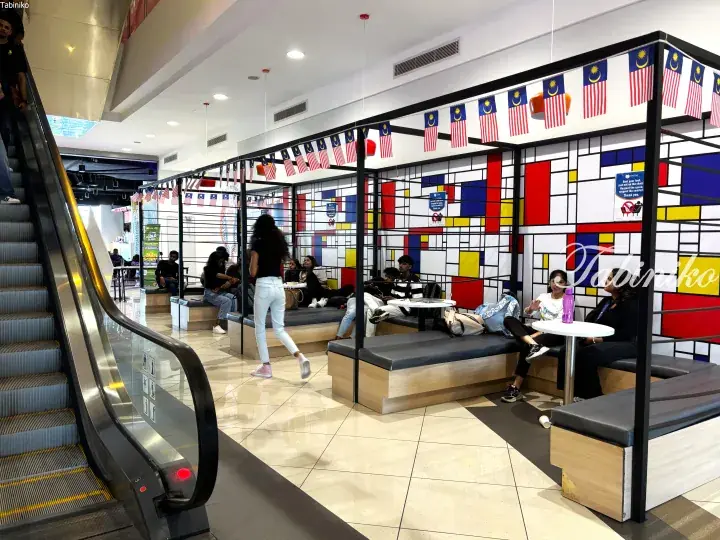 Students relaxing in a modern campus lounge with Mondrian-style decor and Malaysian flags near an escalator.