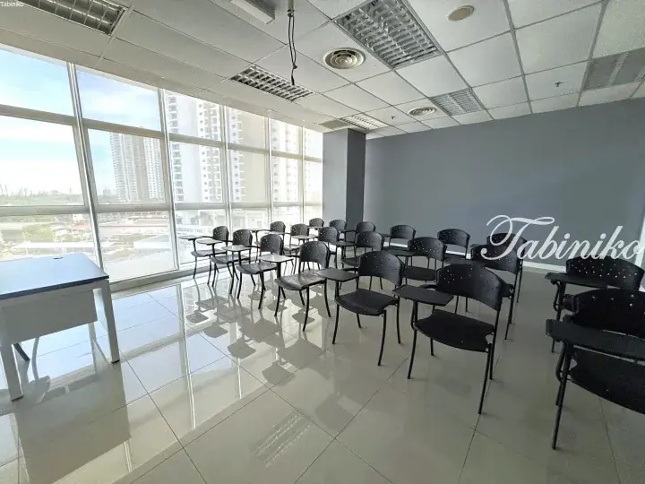 Bright classroom with black chairs and white desk near large windows overlooking city buildings.